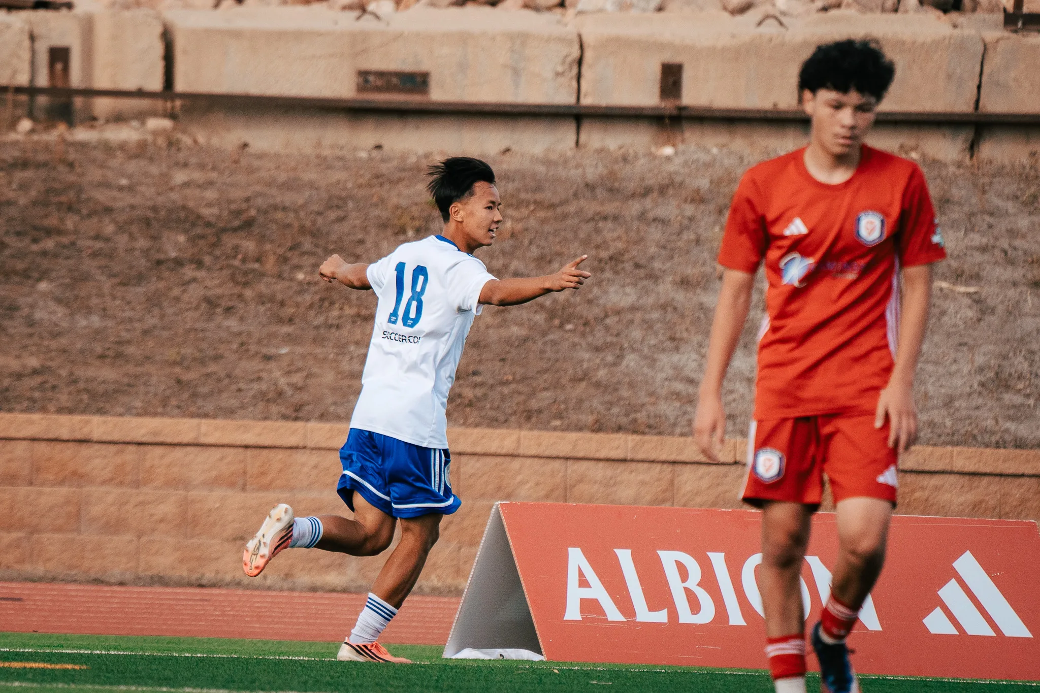 Albion SC Colorado number 18 runs off smiling and pointing as a blurred AC River player walks past in the foreground