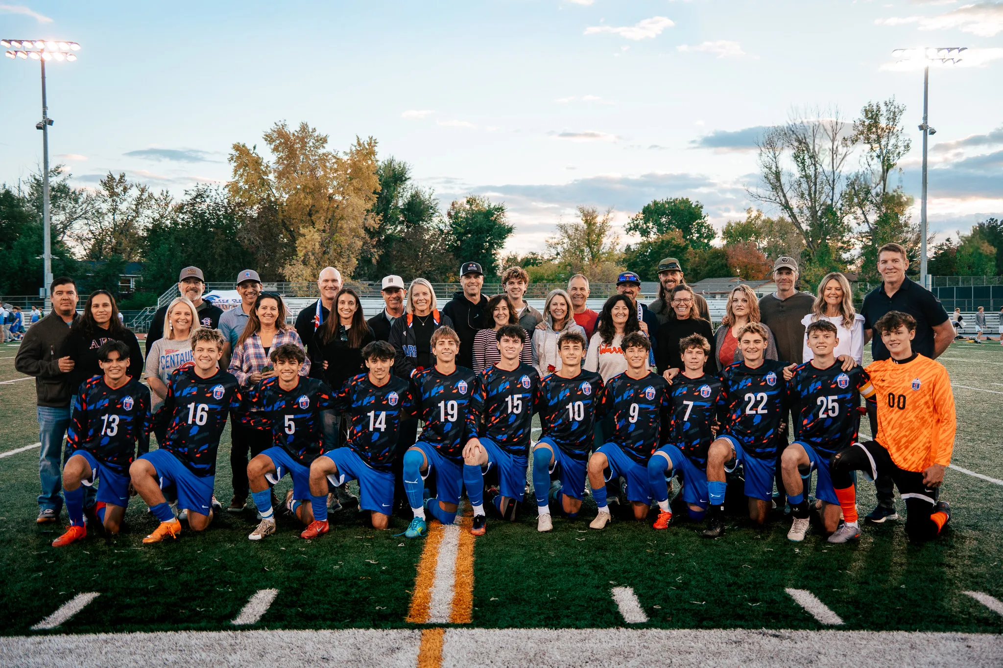Twelve Centaurus seniors kneel in a row in dark patterned jerseys, the goalkeeper in orange at the right end, with their families standing close behind them under the stadium lights at dusk