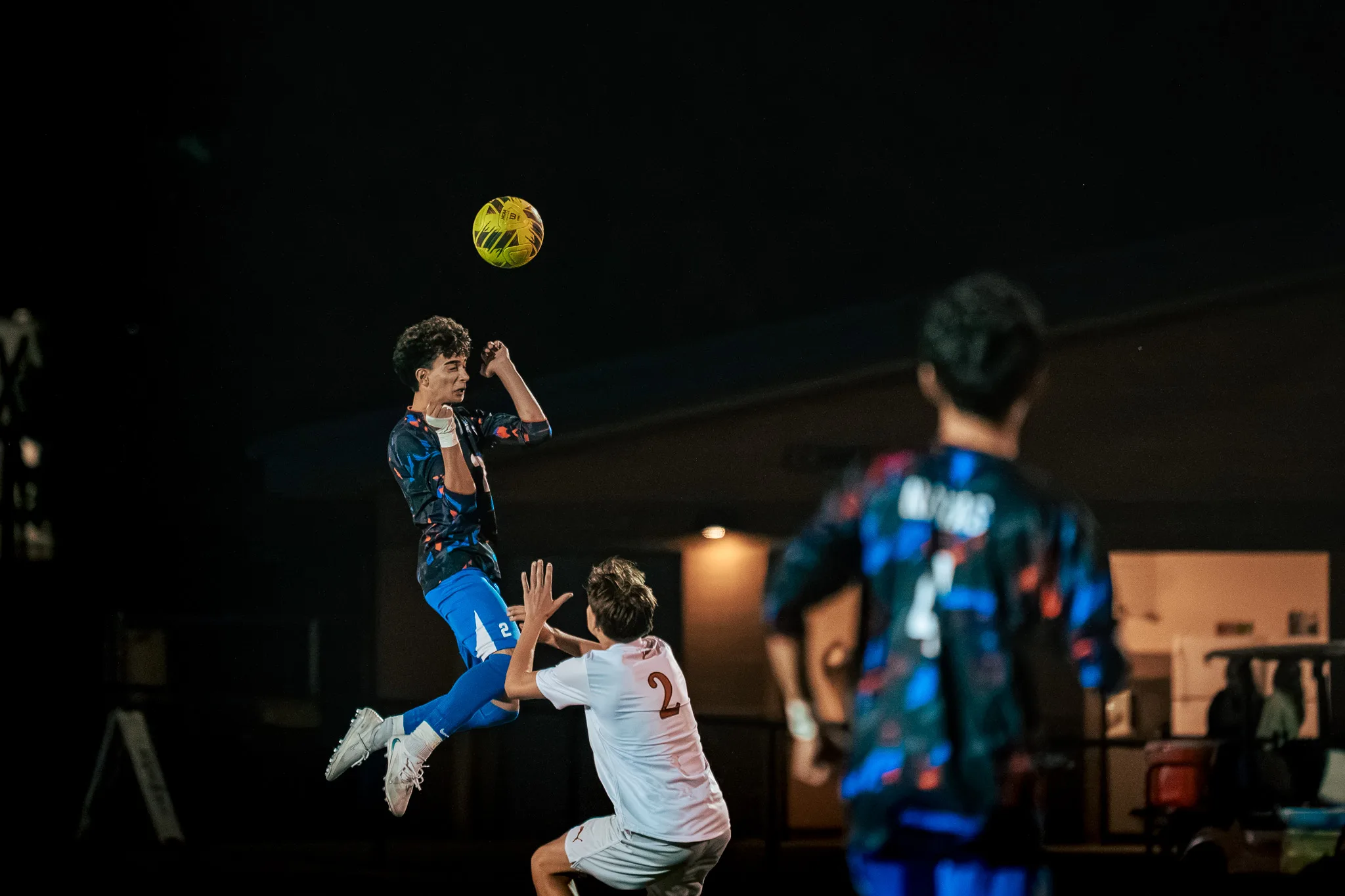 Centaurus number 2 leaps to head the ball over Mead number 2 at night as a blurred teammate watches from the foreground
