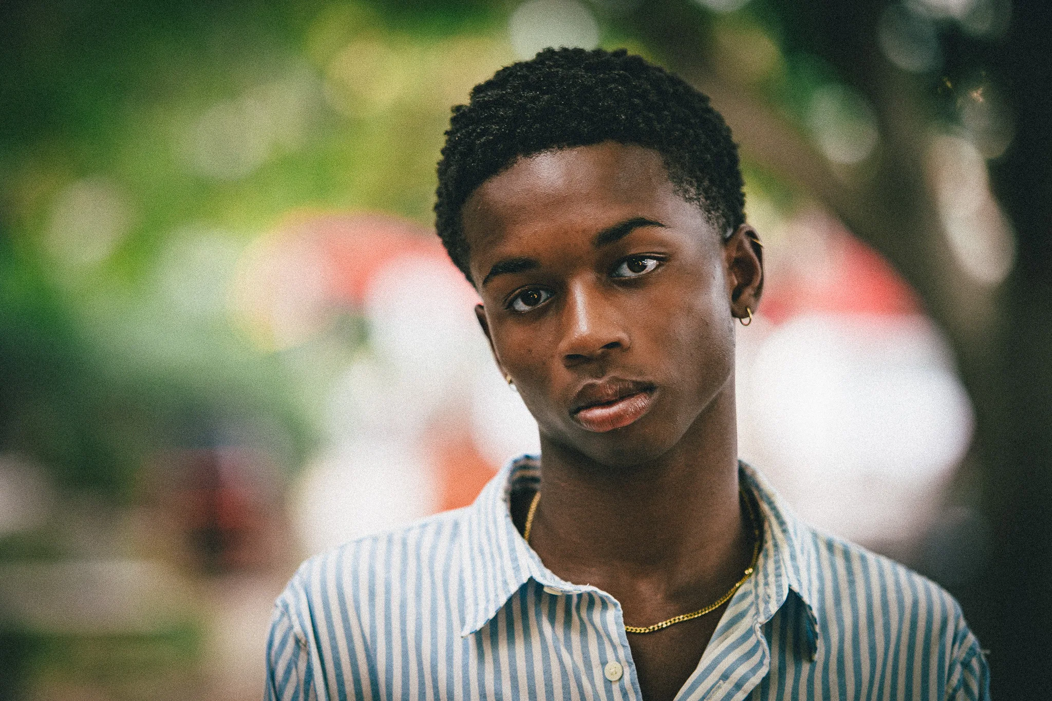 Charlie, in a blue striped shirt and gold chain, looks into the camera against soft green and red bokeh.